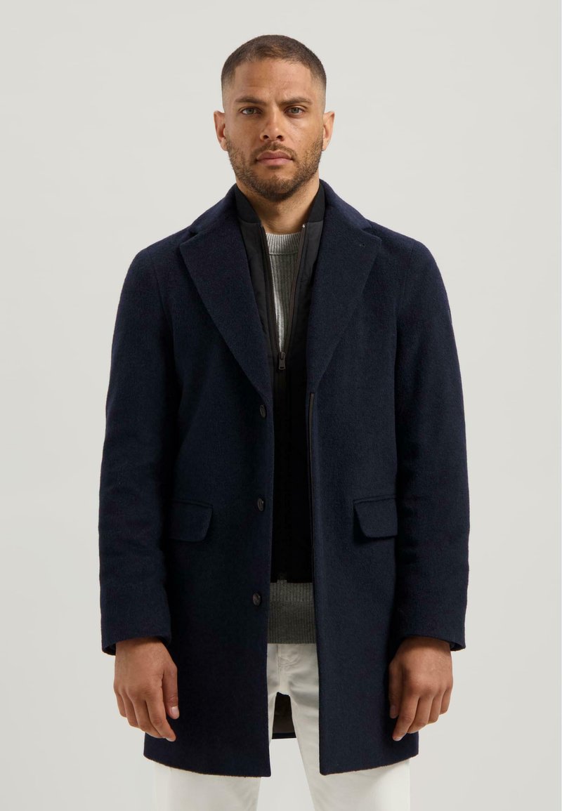 Marineblauwe wollen jas met notch-lapels, enkelvoudige sluiting en twee zijzakken, gelaagd over een gestructureerde grijze trui. Indrukwekkende silhouet.