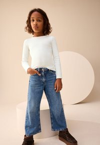 Haut blanc à manches longues, jean large en denim bleu, et bottes imprimé léopard marron ; avec un fond circulaire proéminent.