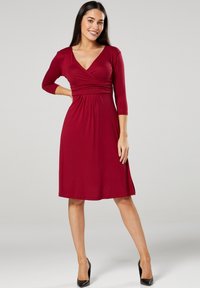 Burgunderfarbenes V-Ausschnitt-Kleid mit drei Viertelärmeln, tailliertem Oberteil und fließendem Rock. Glatter Stoff mit Faltendetails. Kombiniert mit schwarzen Absätzen.