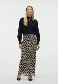 Egy nő fekete hosszú ujjú blúzban, nyakkendővel és fekete maxi szoknyában van, amely fehér és sárga margaréta mintával díszített.