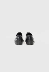 Chaussures en cuir noir avec une finition lisse, bout arrondi et petit talon. Les détails incluent des coutures le long des bords et un design minimaliste.