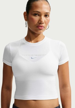 Femme avec des boucles d'oreilles anneaux portant un t-shirt crop top blanc moulant avec le texte en relief "Nike Shox" et un petit logo Nike bleu marine.
