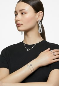 Srebrna biżuteria wyeksponowana, w tym duże kryształowe kolczyki, naszyjnik w kształcie gwiazdy z perłami oraz pierścionki z akcentami kamieni szlachetnych na ciemnej koszuli.