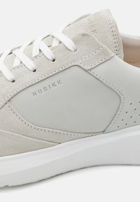 Zapatillas de ante gris claro con cordones blancos. Presentan detalles de cuero suave y un diseño perforado en el lateral. Logo embossado en el lado exterior.