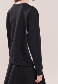 Czarna bluza z okrągłym dekoltem, z długimi rękawami i kontrastowym białym panelem bocznym. Materiał wydaje się gładki i fakturowany.