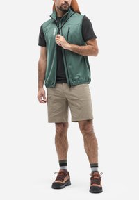 Grüne Zip-Weste mit Netzstruktur, schwarzes Kurzarmshirt, beiger Cargo-Shorts und braune Wanderschuhe mit orangen Akzenten.