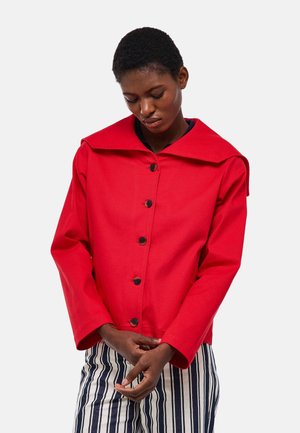 Personne aux cheveux courts portant une veste rouge vif boutonnée et un pantalon à rayures verticales noires et blanches, regardant vers le bas avec les mains jointes.