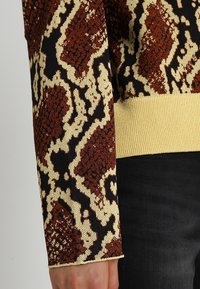 Strickpullover mit gelbem Grund und einem strukturierten, mehrfarbigen Schlangenprint in Braun und Schwarz. Gerippte Ärmelbündchen und Saum in Gelb.