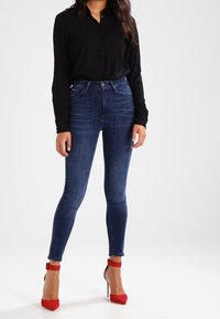 Hoch geschnittene dunkelblaue Skinny-Jeans kombiniert mit einem schwarzen Hemd mit Knöpfen und roten High Heels mit Knöchelriemen.