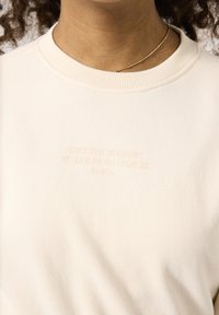 Sudadera de un beige claro con cuello redondo, con texto en relieve: "TOMA EL CAMINO HACIA LUGARES DESCONOCIDOS" en un tono sutil. Tejido suave.