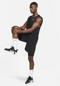 Must värviga sporditop Nike logo ja mustade lühikesed püksid, valged sokid ja mustad tossud. Figuur on venitusasendis.