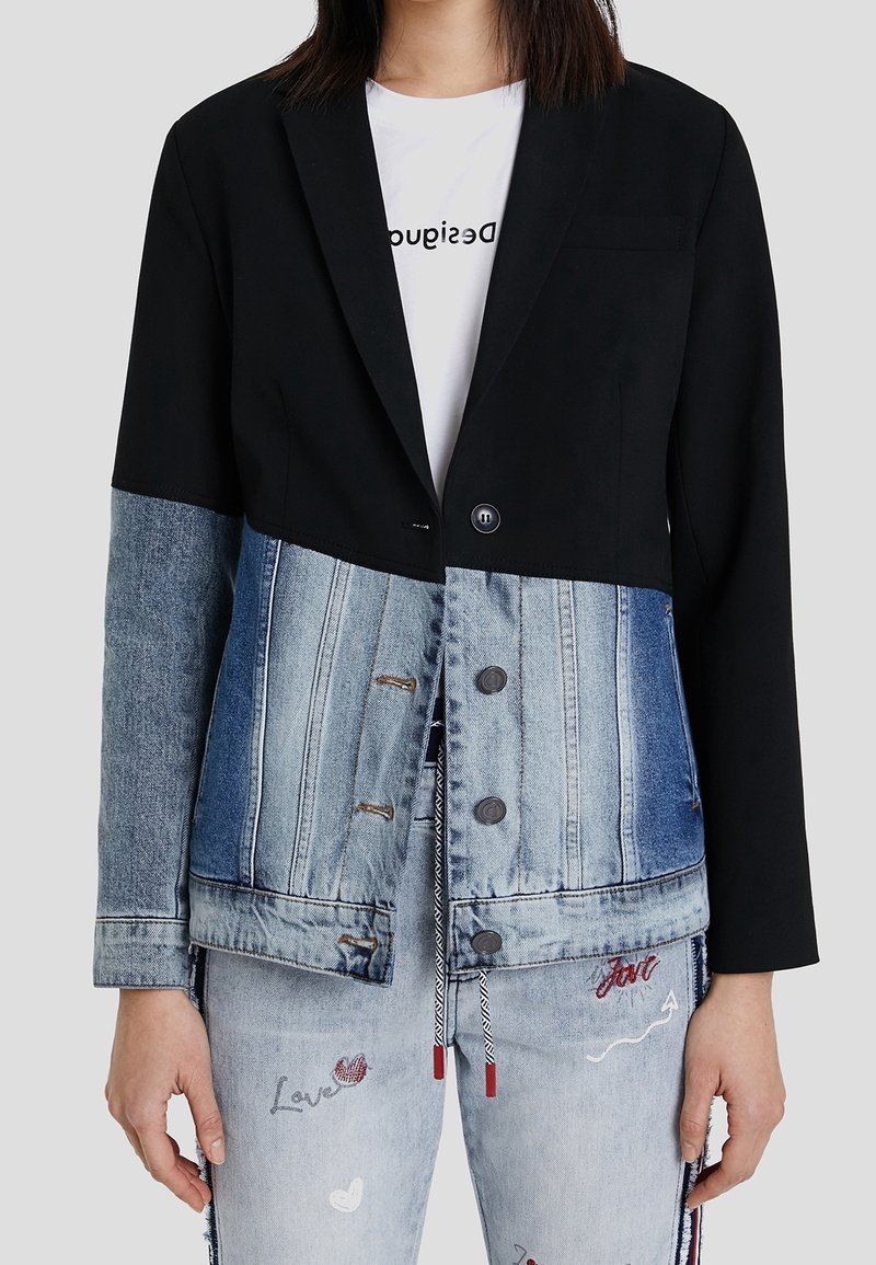 Femme portant un blazer noir combiné avec une veste en jean bleue et un jean bleu clair avec des motifs brodés et du texte.