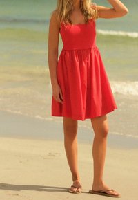 Robe sans manches rouge en forme de A avec un décolleté carré, taille froncée et tissu lisse. Associée à des tongs marron sur une plage.