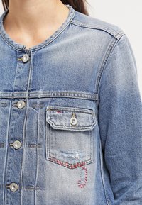 Jeansjacka i ljusblått, med metallknappar, två bröstfickor, blekta områden och röd broderad detalj på en ficka.