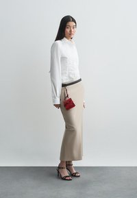 Γυναίκα ντυμένη με λευκό πουκάμισο με κουμπιά, μπεζ μακρύ φούστα, μαύρες γόβες και μικρή κόκκινη τσάντα που κρέμεται από μαύρη ζώνη, standing σε γκρι δάπεδο.