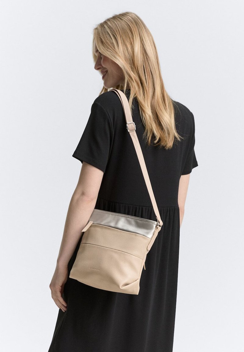 Beige Leder Umhängetasche mit einem silbernen Akzentpaneel, ausgestattet mit einem Reißverschluss und einem verstellbaren Riemen, gehalten von einer Frau in einem schwarzen Kleid.