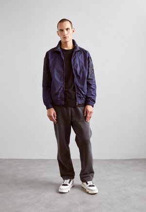 Marineblaue Nylonjacke mit Reißverschluss, gerippten Bündchen und gemusterten Ärmeln, kombiniert mit einem schwarzen T-Shirt und grauen Hosen, getragen mit weißen Sneakers.