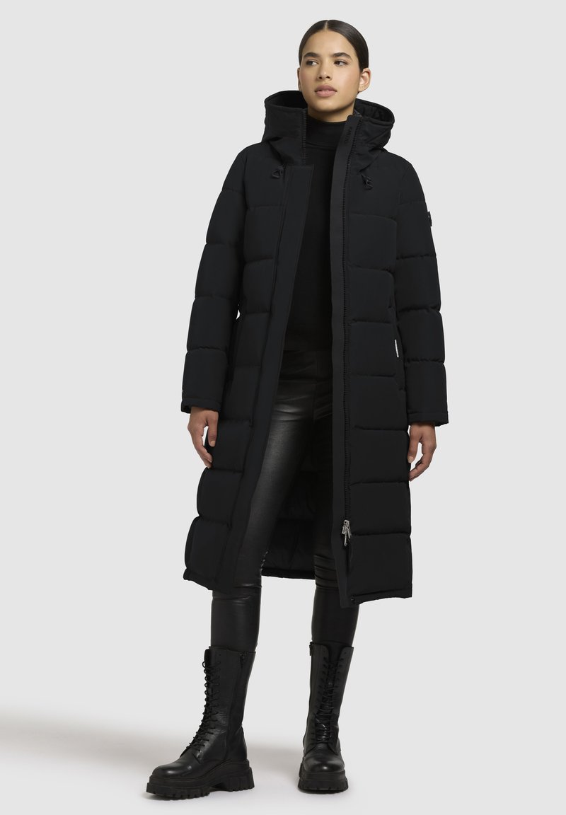 Cappotto imbottito nero con design lungo e trapuntato, cappuccio e chiusura con zip. Abbinato a pantaloni di pelle nera e stivali da combattimento con lacci.