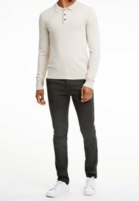 Beżowa polo z długim rękawem, z kołnierzykiem i trzema ciemnymi guzikami, zestawiona z ciemnymi dżinsami slim-fit i białymi trampkami.