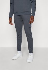 Mann som har på seg mørkegrå joggebukser med sidelommer, kombinert med hvite joggesko og hvite sokker mot en ensfarget bakgrunn.