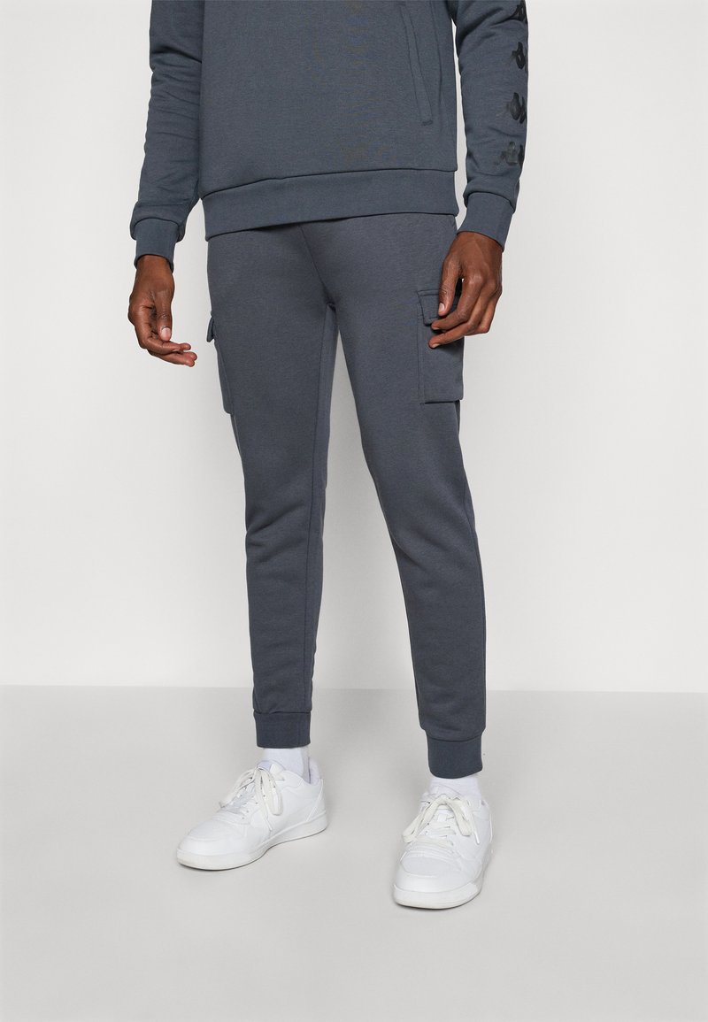 Mann som har på seg mørkegrå joggebukser med sidelommer, kombinert med hvite joggesko og hvite sokker mot en ensfarget bakgrunn.