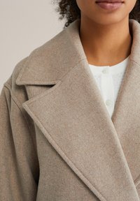 Bež plašč iz volna z ovratnikom z režo in oblikovanimi rameni. Vidni šivi in mehka tekstura, slojevito oblečen preko belega zgornjega dela s gumbi.