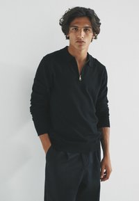 Černý pulovr s čtvrťovým zipem a límcem. Měkká látka, přiléhavý střih a žebrované manžety. Model stojí před bílým pozadím.