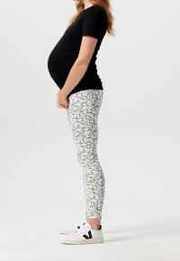 Strój ciążowy składający się z czarnej bluzki z krótkim rękawem w połączeniu z legginsami w kwiatowy wzór w jasnym zielonym kolorze, podkreślonym czarnymi i białymi detalami.