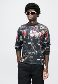 Μπλούζα με μακρύ μανίκι που διαθέτει κολάζ με εικόνες μπάσκετ, κυρίως σε μαύρο χρώμα με πολύχρωμες λεπτομέρειες και υφή υφάσματος. Μαύρο παντελόνι ολοκληρώνει την εμφάνιση.