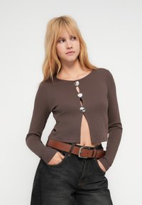 Νεαρή γυναίκα με ξανθά μαλλιά φοράει ένα καφέ κοντό τοπ με τρία ασημένια κουμπιά και μαύρο τζιν ψηλόμεσο με καφέ ζώνη.