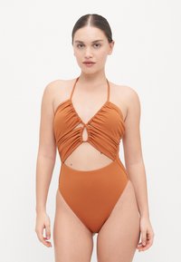 Maillot de bain une pièce marron avec des détails froncés et des sections découpées. Il dispose de fines bretelles et d'un design à jambe haute pour une coupe ajustée.