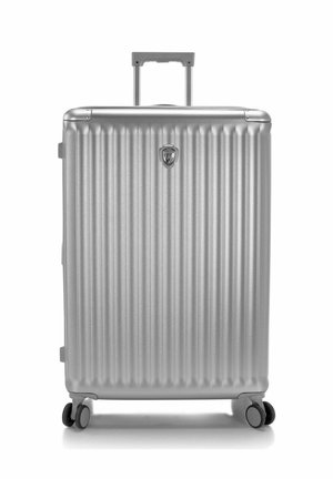 Valise rigide argentée avec rainures verticales, poignée télescopique sortie et quatre roulettes noires pivotantes sur fond blanc.