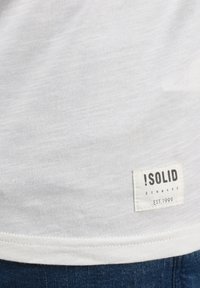 Balts tekstila T-krekls ar teksturētu audumu, noapaļotu apakšmalu un redzamu etiķeti ar detaļām "!SOLID", "DANAMA", un "EST. 1999".