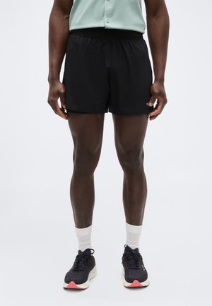 Άνδρας που φοράει μαύρο αθλητικό σορτς, λευκές κάλτσες μέχρι τον αστράγαλο και μαύρα παπούτσια τρεξίματος, στέκεται μπροστά σε απλό φόντο.