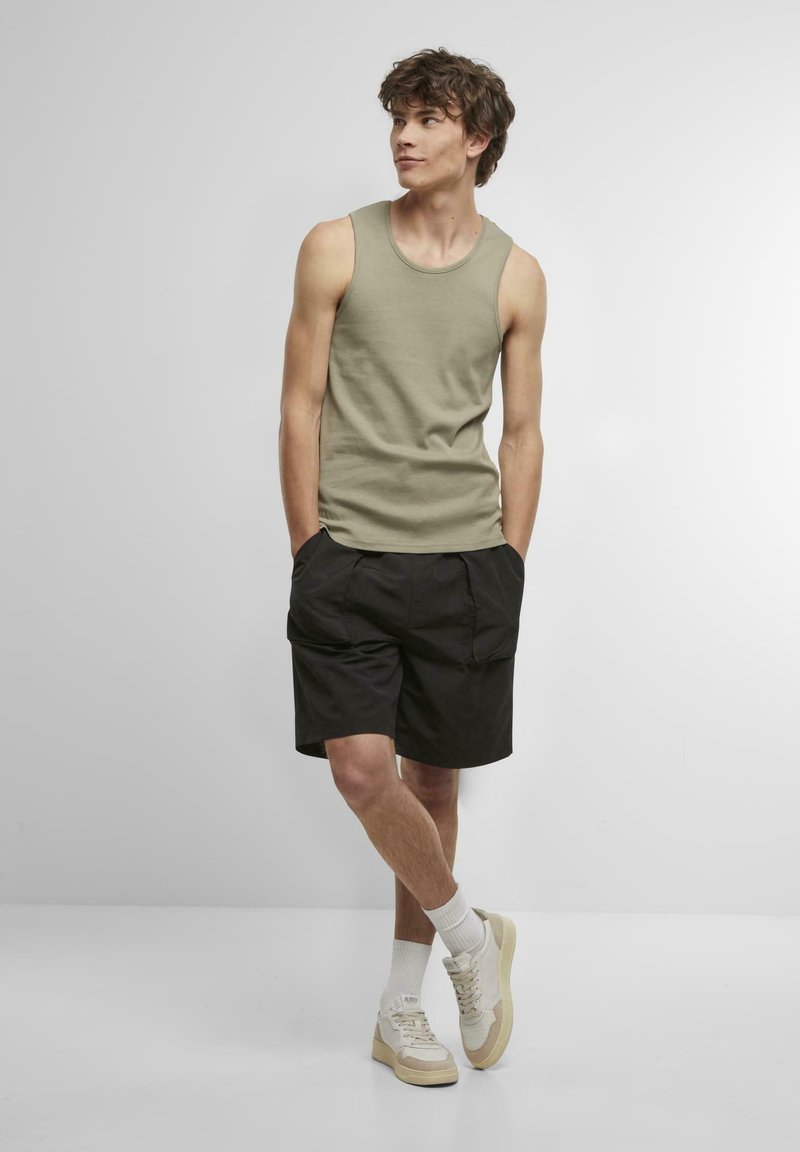 Olivgrünes Trägershirt, schwarze Shorts und cremefarbene Sneakers. Model steht mit den Händen in den Taschen; athletische Figur, lässiger Look. Weicher Stoff.