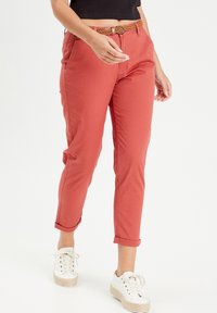 Personne portant un pantalon court corail avec une ceinture tressée et des baskets blanches, debout devant un fond blanc uni.