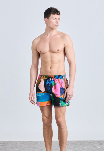 Άνδρας χωρίς μπλούζα, όρθιος, φοράει πολύχρωμο μαγιό με αφηρημένο μοτίβο, κοιτώντας προς τα αριστερά του, σε απλό φωτεινό γκρι φόντο.