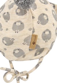 Beige knitted baby hat with gray sheep pattern, a tied chin strap, a pom-pom on top, and a small brown stitched brand label.