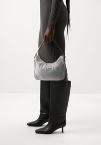 Personne vêtue d'une robe sombre côtelée et de bottes noires montantes tenant un petit sac à main argenté avec une poignée supérieure et une plaque de logo.