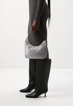 Osoba v tmavých žebrovaných šatech a černých kozačkách ke kolenům drží malou stříbrnou kabelku s horním držadlem a logem.