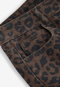 Tissu en denim marron avec un motif léopard noir, comprenant une poche avant et des détails en métal sur les boutons. Surface texturée avec des coutures visibles.