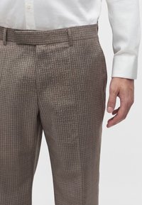 Homme portant un pantalon habillé marron à carreaux et une chemise blanche boutonnée, la main droite détendue le long du corps.