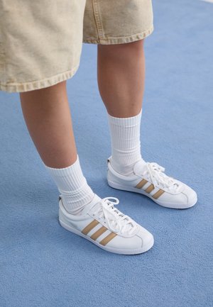 Beine, die beige Shorts, weiße Sportsocken und weiße Sneakers mit hellbraunen Streifen tragen, stehen auf blauem Boden.