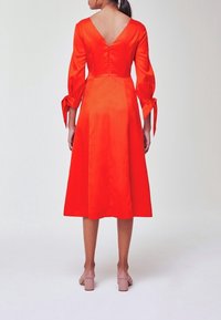 Femme portant une robe midi rouge vif à manches longues avec un décolleté dos en V profond et des poignets de manches noués, associée à des sandales à talons blocs couleur nude.