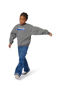 Sudadera gris de cuello redondo con puños acanalados que presenta un logotipo gráfico en azul y blanco, combinada con pantalones cargo de denim de pierna ancha y zapatillas blancas.