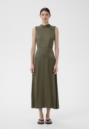 Kobieta stojąca w oliwkowej sukience midi bez rękawów z wysokim kołnierzem i czarnych płaskich sandałach na jednolitym białym tle.