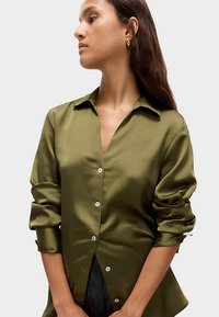 Olive green satin shirt with a collared design, front buttons, and rolled sleeves. Textured fabric reflects light.