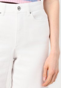 Bílé džíny s klasickým pětikapsovým designem, vybavené zipem a knoflíkem. Hladká textura s jemnými švy.