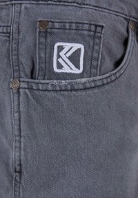 Hallharjane denimkangas, millel on tagatasku metallist studide ja valge tikandiga logopatch, näidates struktureeritud disaini ja sujuvat tekstuuri.