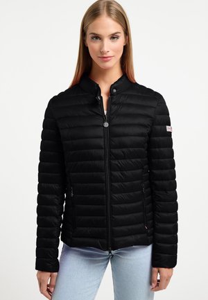 Kobieta w czarnej, pikowanej kurtce zapinanej na suwak z bocznymi kieszeniami i jasnoniebieskich jeansach, stojąca na jednolitym białym tle.